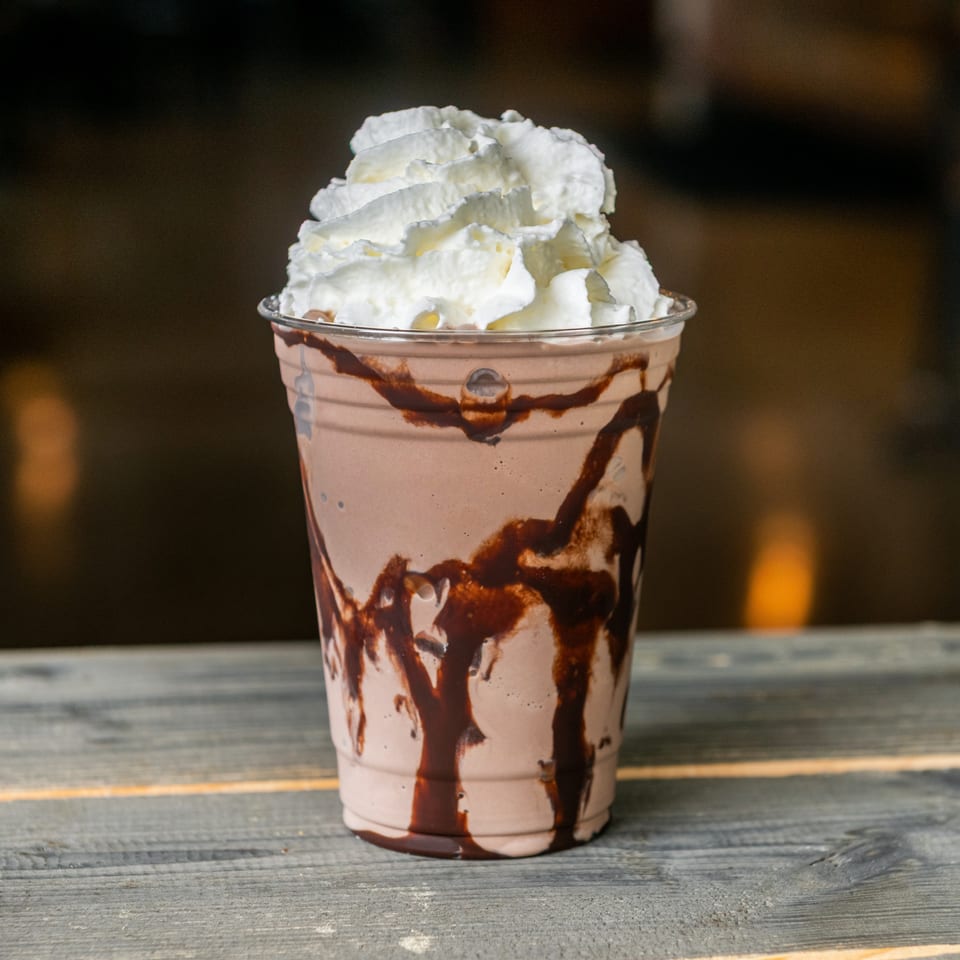Chocolate milkshake drizzled with chocolate sauce and topped with whipped cream, served in a clear cup on a wooden table.