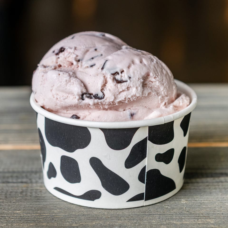 Scoop of pink chocolate-chip ice cream in a cow-print cup on a wooden table.